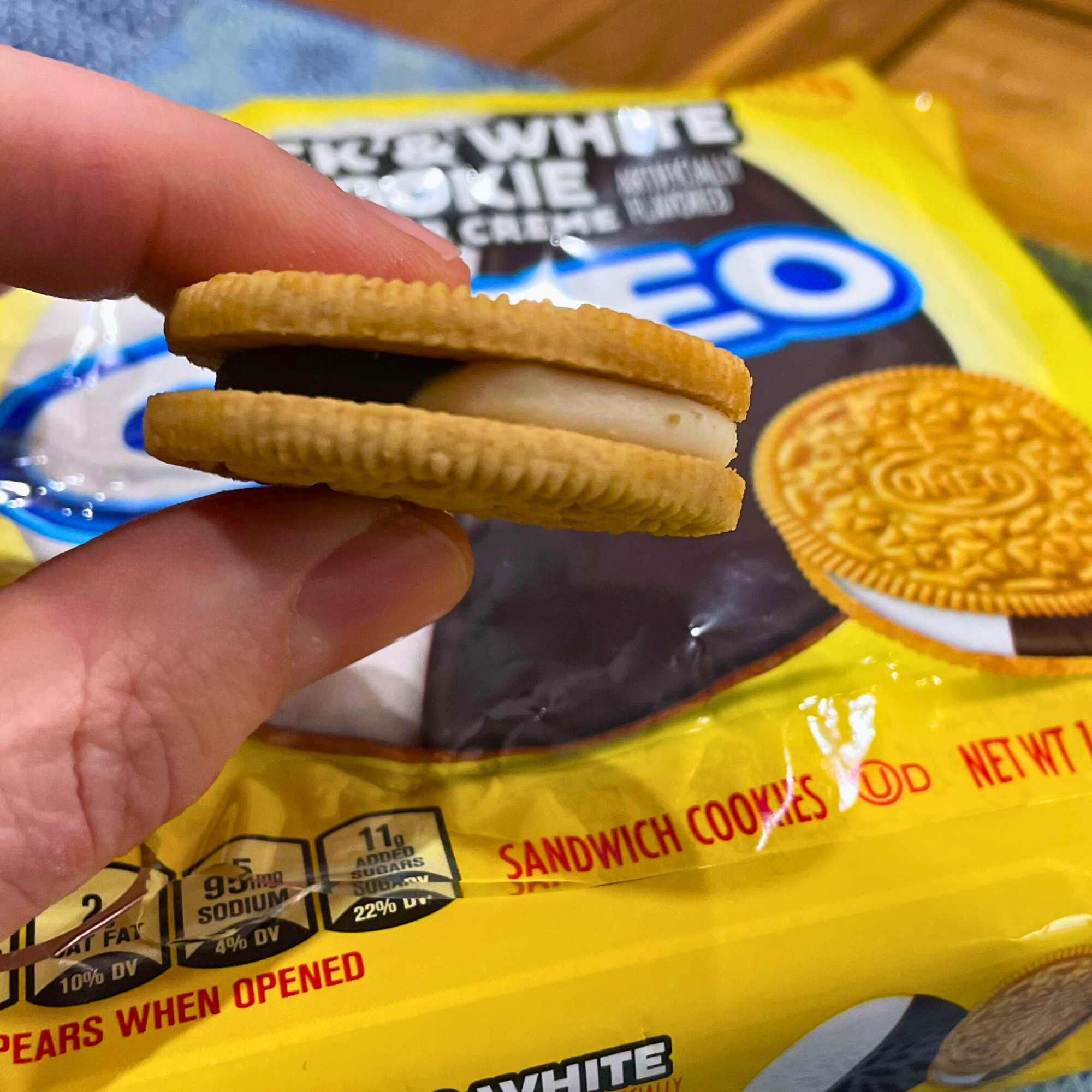 Hand holding a Black & White Cookie Oreo, showing layered crème inside over branded packaging.