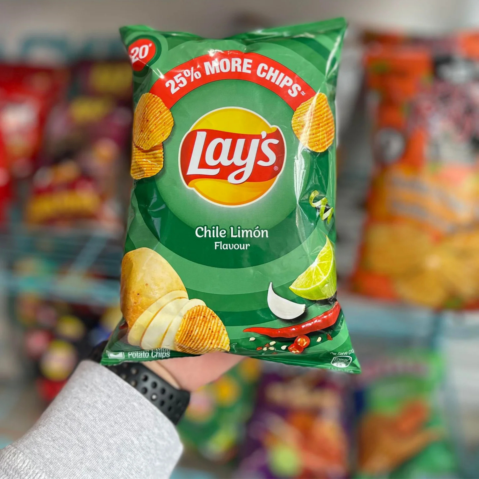 A hand holding a green bag of Lay's Chile Limón flavored chips, labeled “25% More Chips.” The background shows colorful, blurred chip bags.