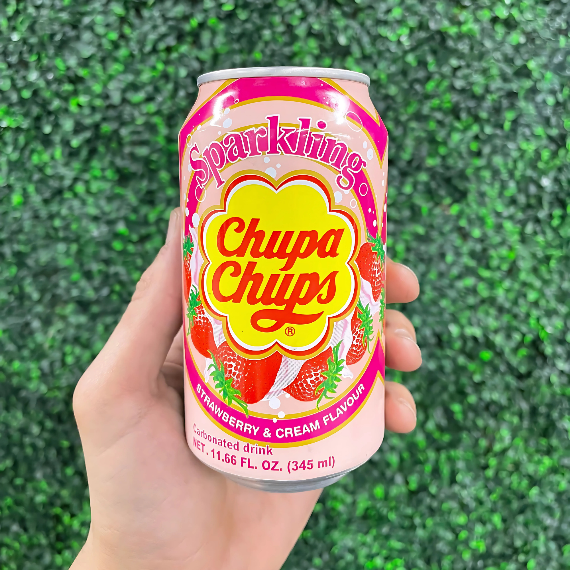 A hand holds a pink can of Chupa Chups sparkling soda, flavored with strawberry and cream. The background is a vibrant green leafy texture.