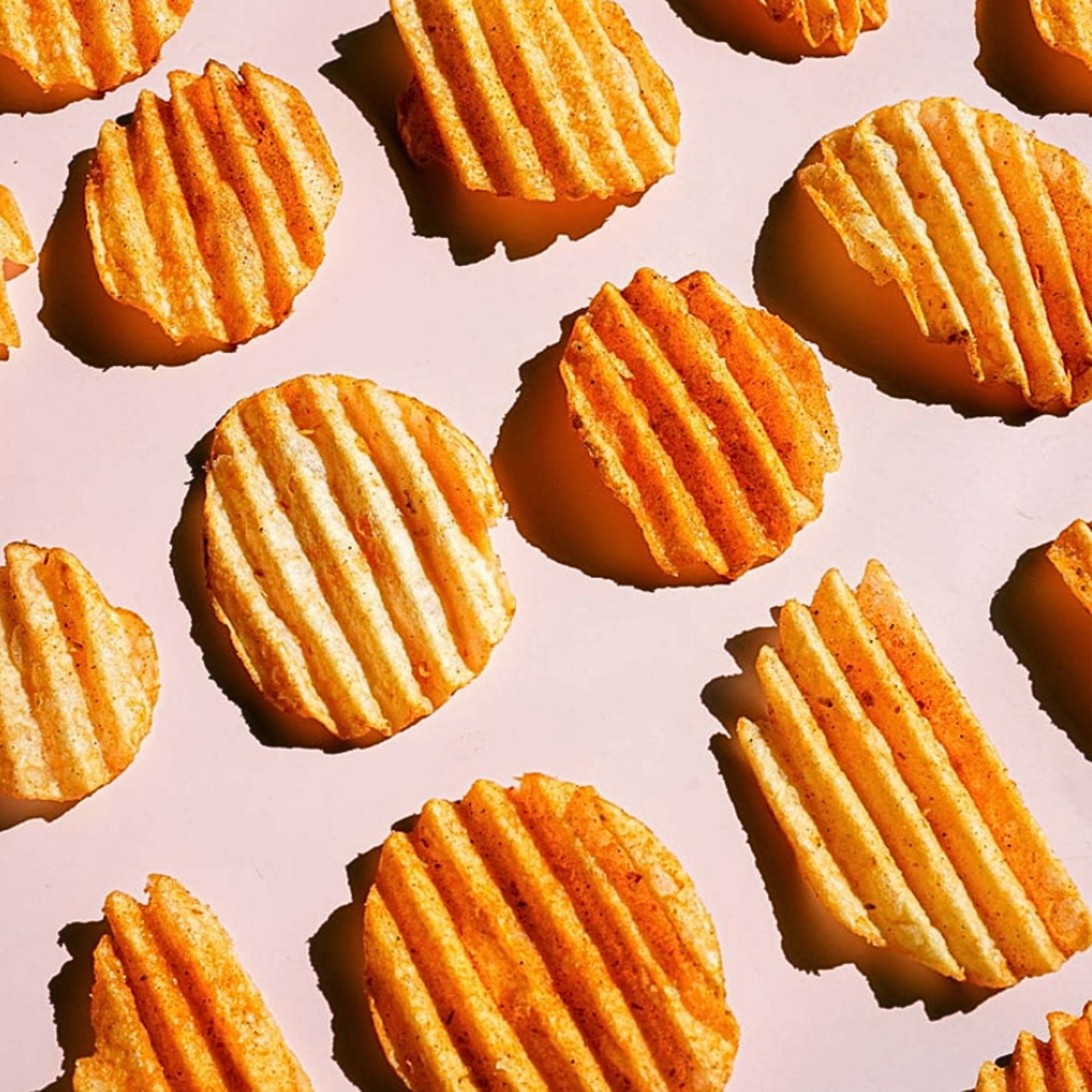 Flat lay of individual ridged Lay’s chips arranged evenly on a light pink surface, showcasing chip thickness and seasoning.