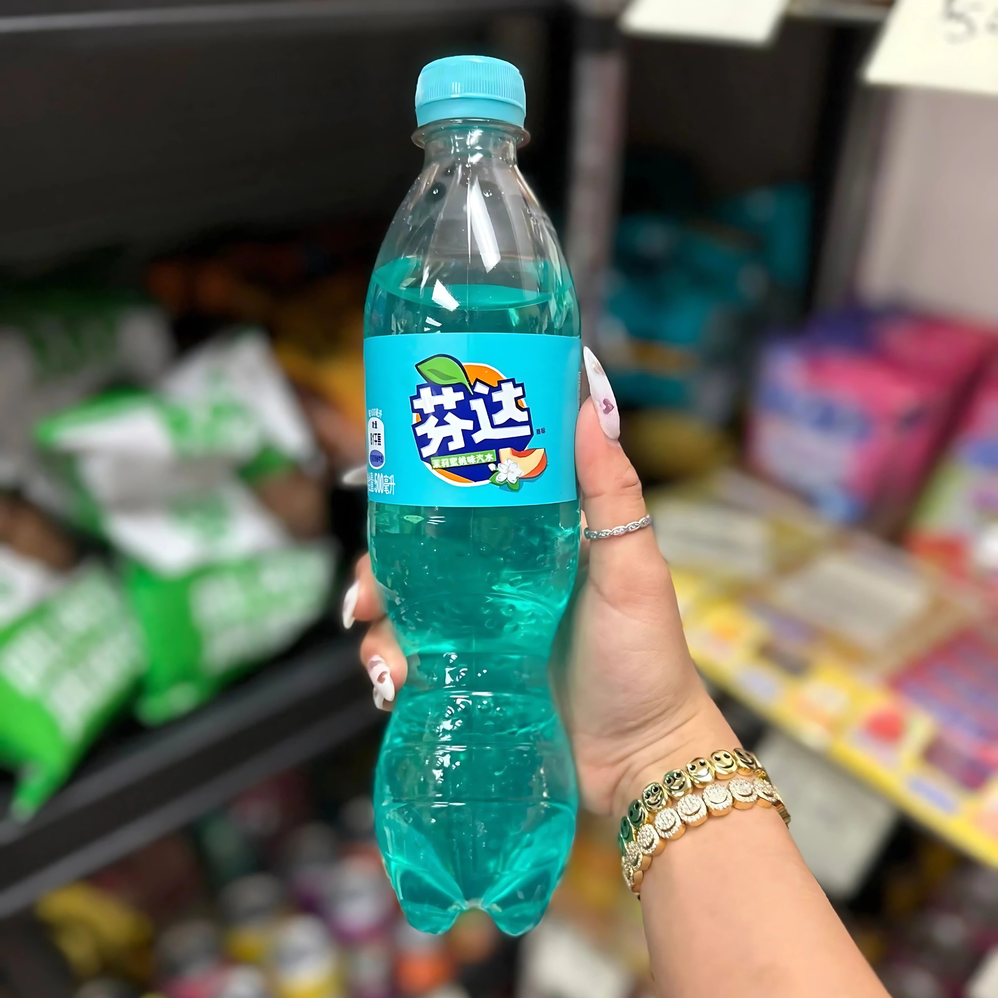 A hand with manicured nails and gold bracelets holds a bottle of Jasmine Peach Fanta in front of a store shelf.