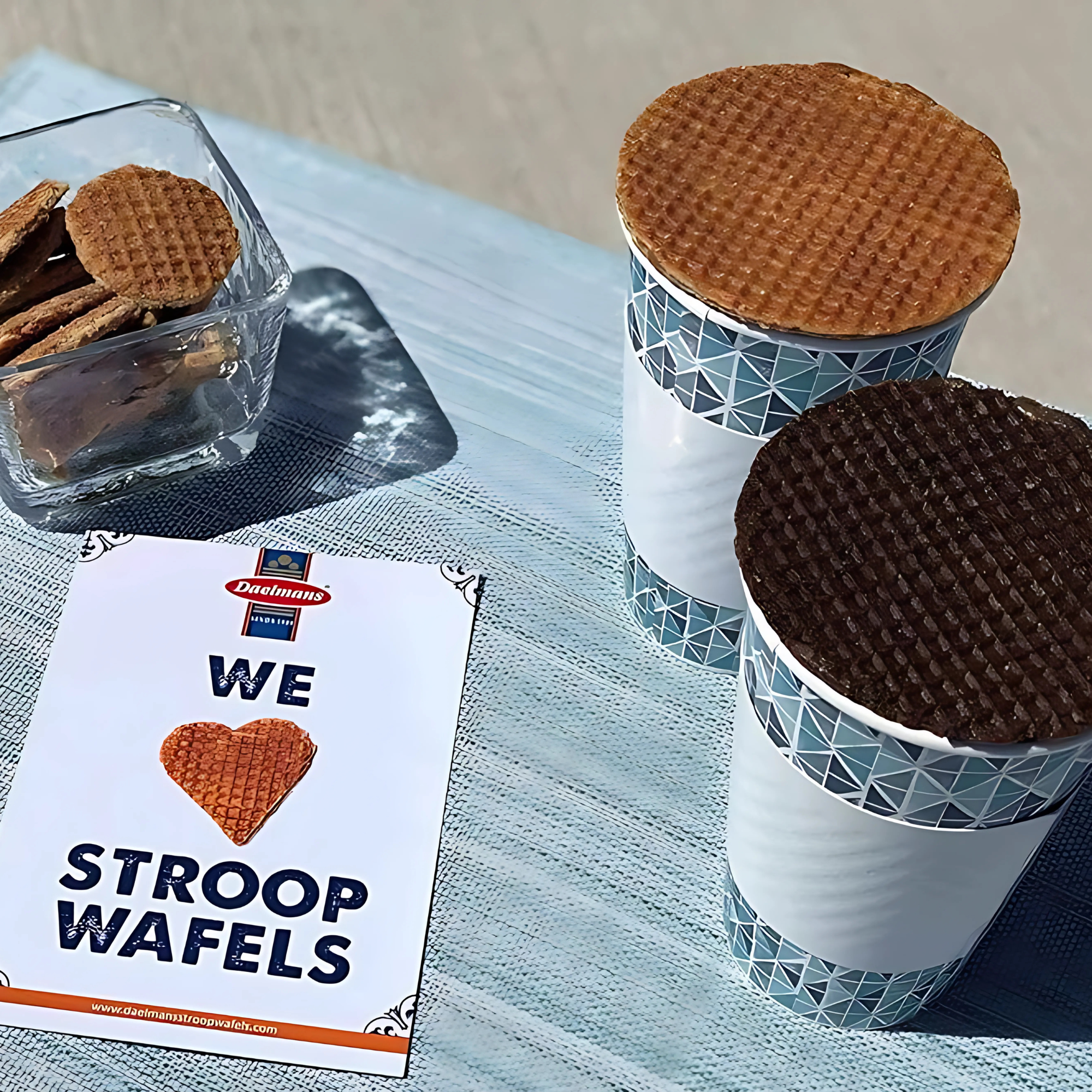 Two cups with stroopwafels on top sit on a blue table. A glass bowl holds more stroopwafels. A sign reads "We Love Stroopwafels."