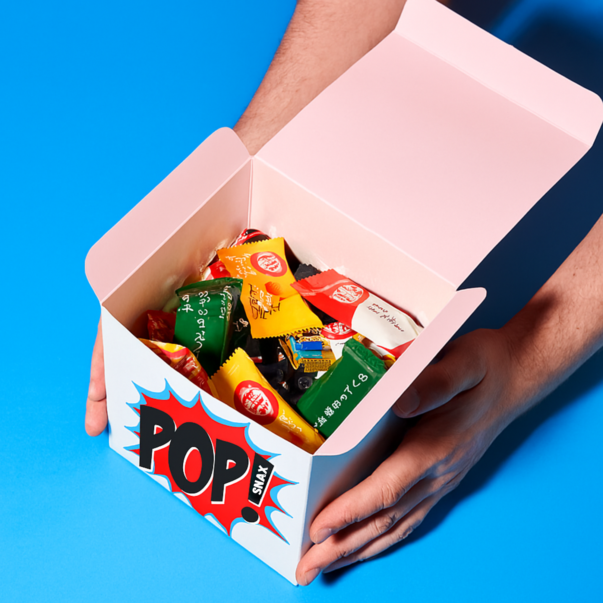 Pop Snax white box filled with assorted Japanese KitKats in white packaging, held by hand against a bold blue background Tokyo Fun Box product shot.