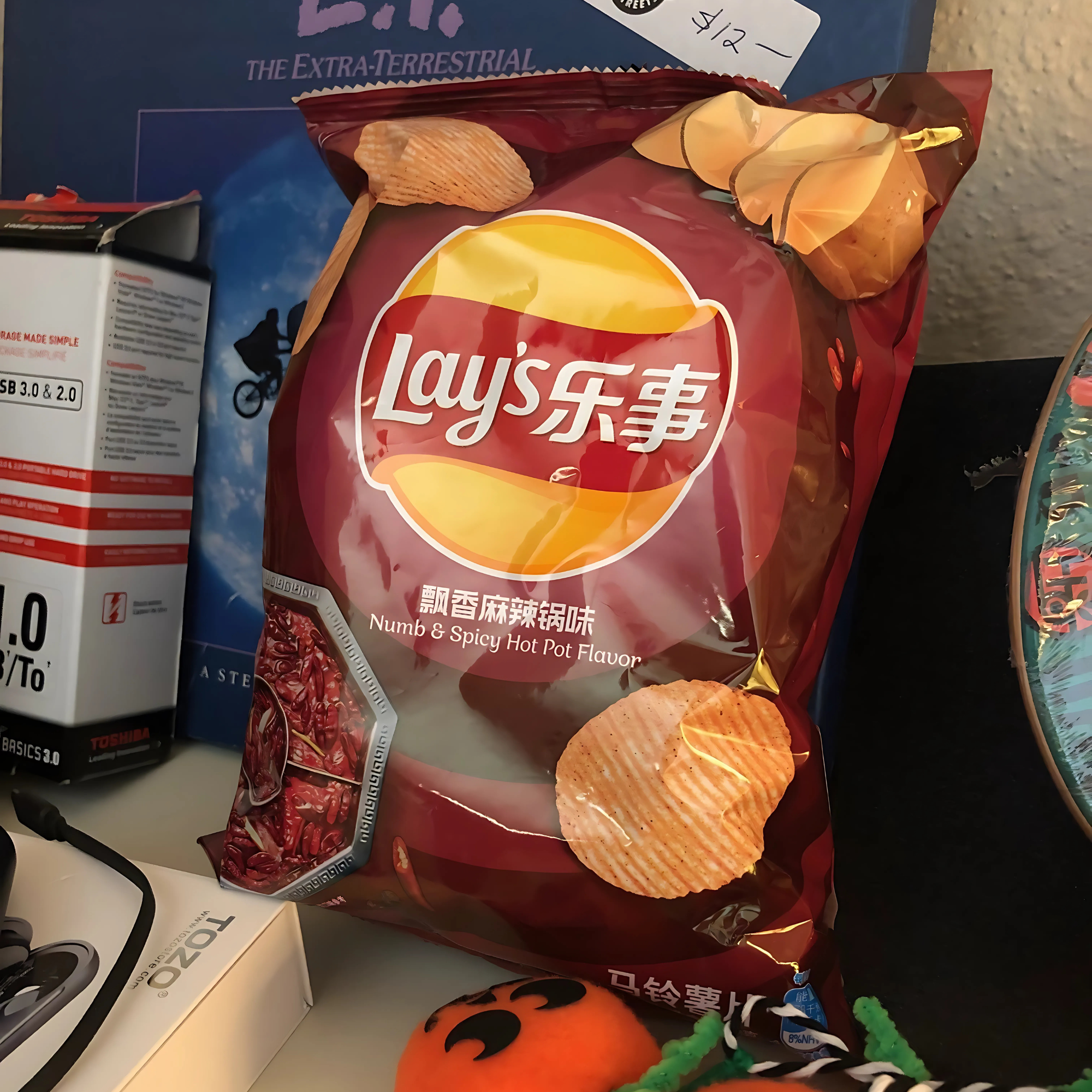 A bag of Lay's Numb & Spicy Hot Pot flavored chips with red packaging is on a cluttered shelf. Nearby are a DVD, a USB box, and a small orange pumpkin.
