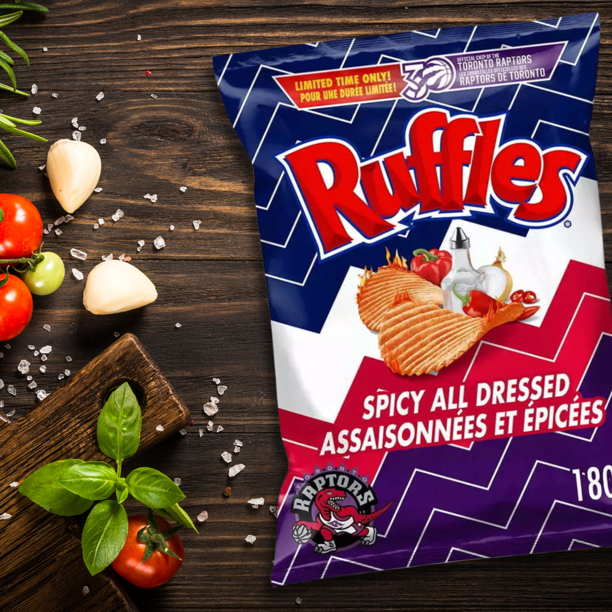 Bag of Ruffles Spicy All Dressed chips with bold colors and Toronto Raptors logo on a wooden table surrounded by fresh tomatoes, garlic, herbs, and salt