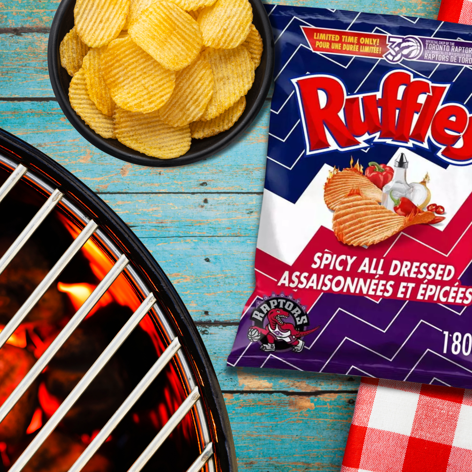 A bag of Ruffles Spicy All Dressed chips with Toronto Raptors branding is on a blue wooden surface. A bowl of chips and a flaming grill are nearby