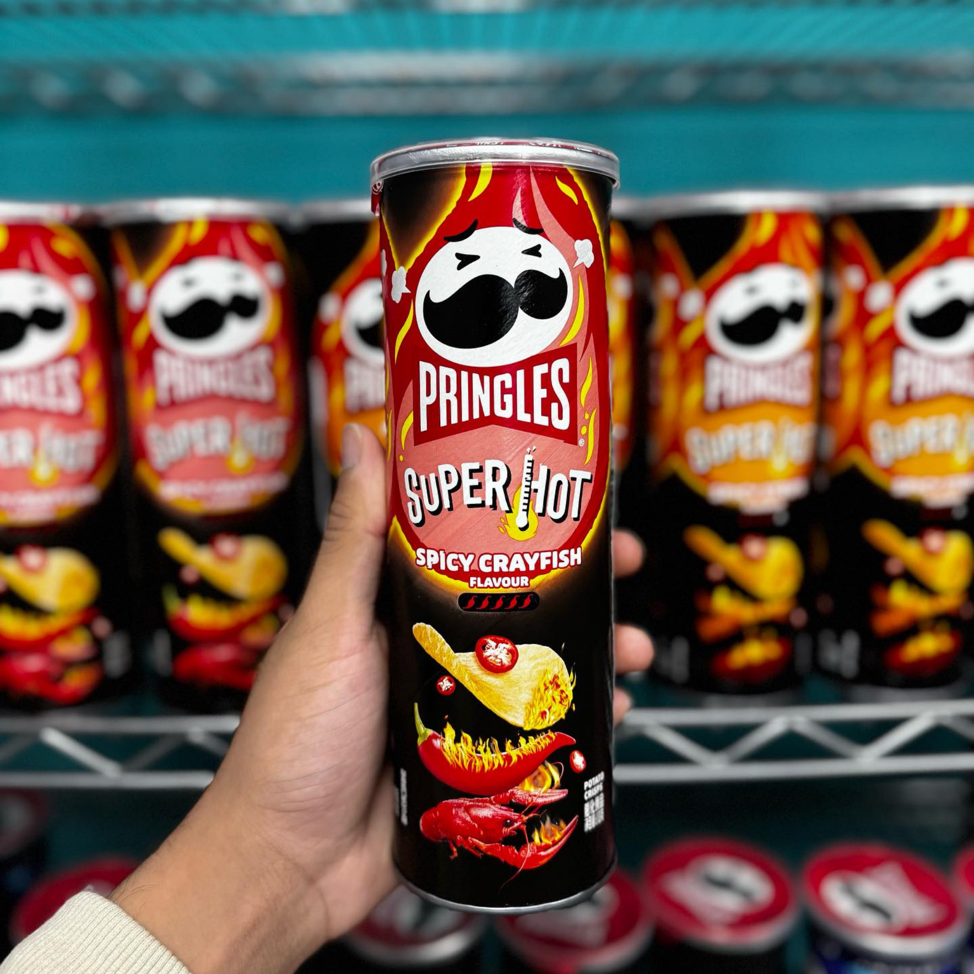 Close-up of a hand holding a Pringles Super Hot Spicy Crayfish flavor can. The fiery design and flames emphasize intense spiciness.