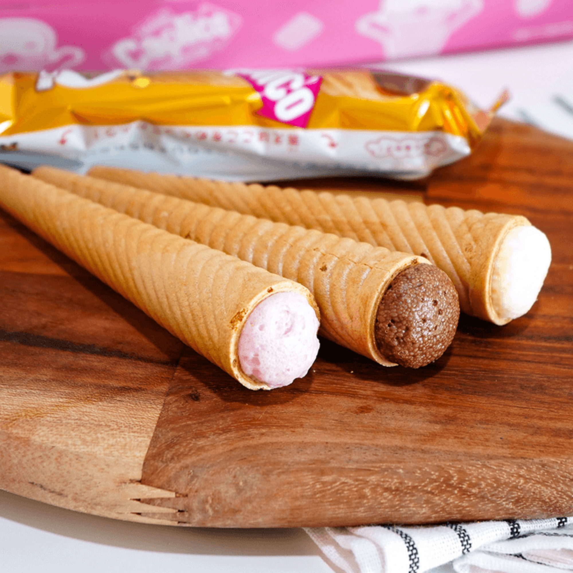 Three Caplico Mini wafer cones from Japan with strawberry, chocolate, and vanilla fillings are displayed on a wooden board. A colorful package lies in the background. Available to buy online.