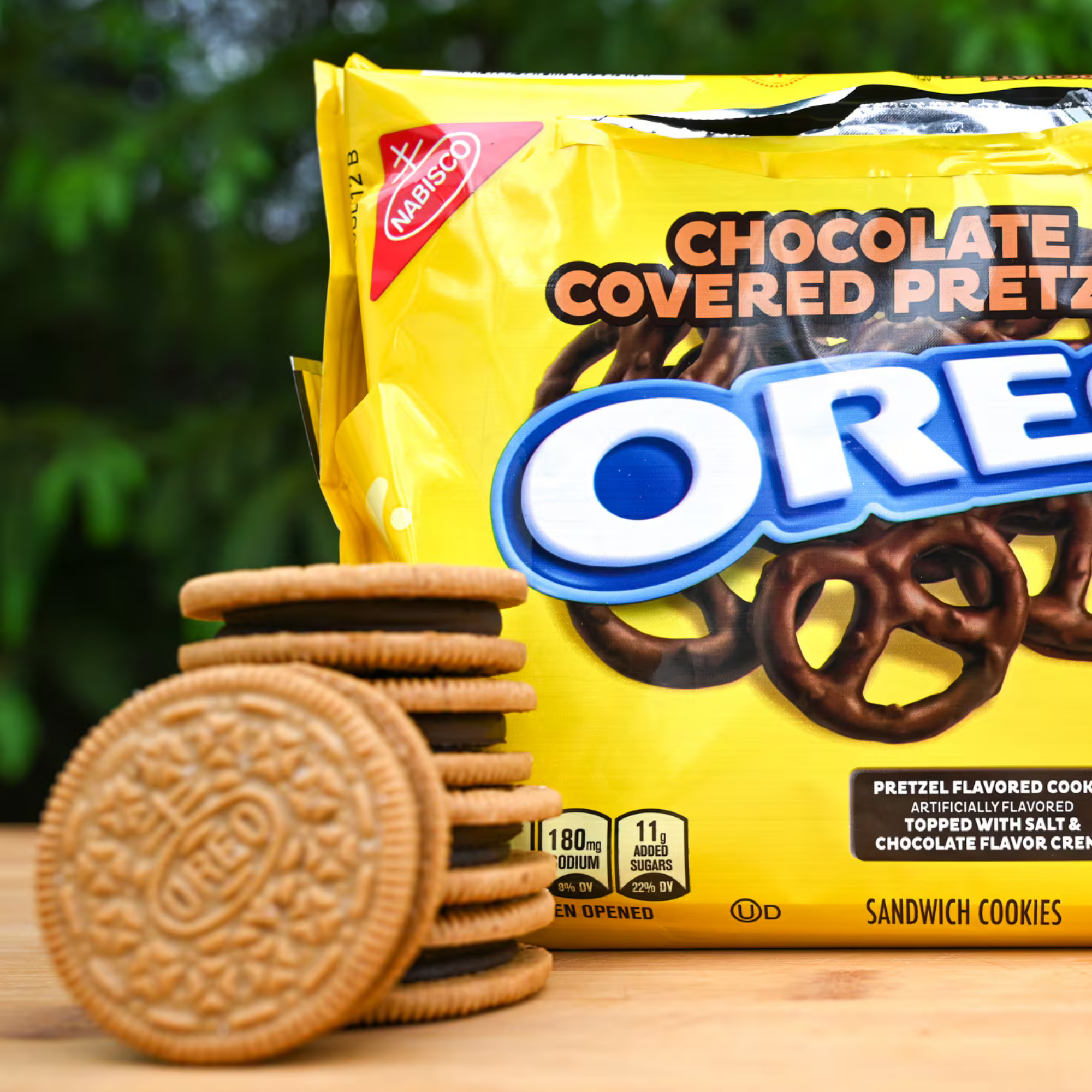 A stack of golden Oreo cookies is in front of a bright yellow package labeled "Chocolate Covered Pretzels Oreo." The scene is outdoors on a wooden table.