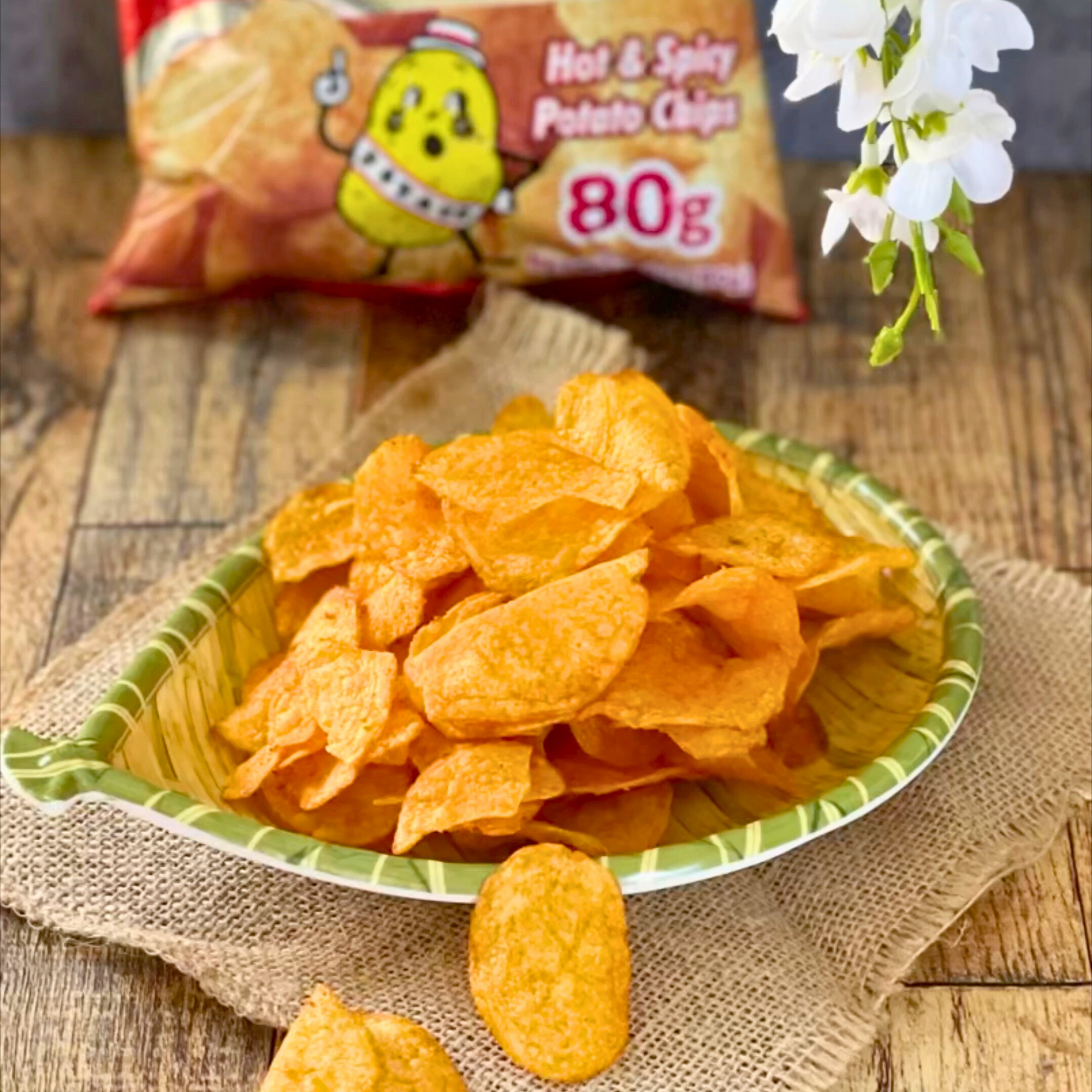 A pile of Calbee hot and spicy potato chips from Japan on a leaf-shaped plate sits on burlap on a wooden table. The chip bag and white flowers are in the background.