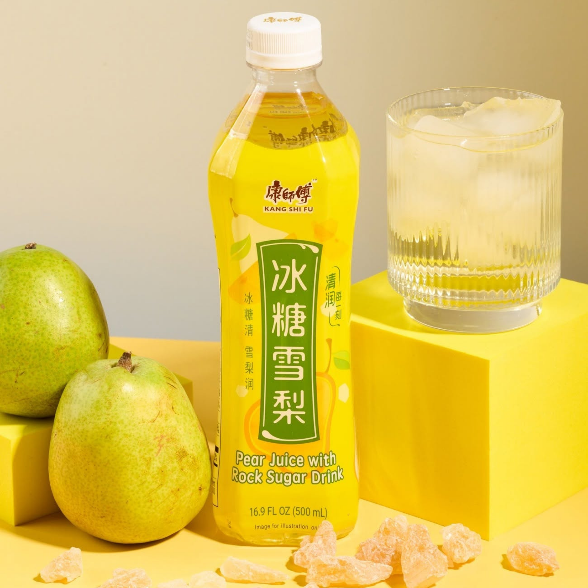 A yellow bottle of Kang Pear Juice from China on a yellow surface, surrounded by fresh green pears and rock sugar chunks. A glass with ice and the juice sits beside it. Available to buy online.