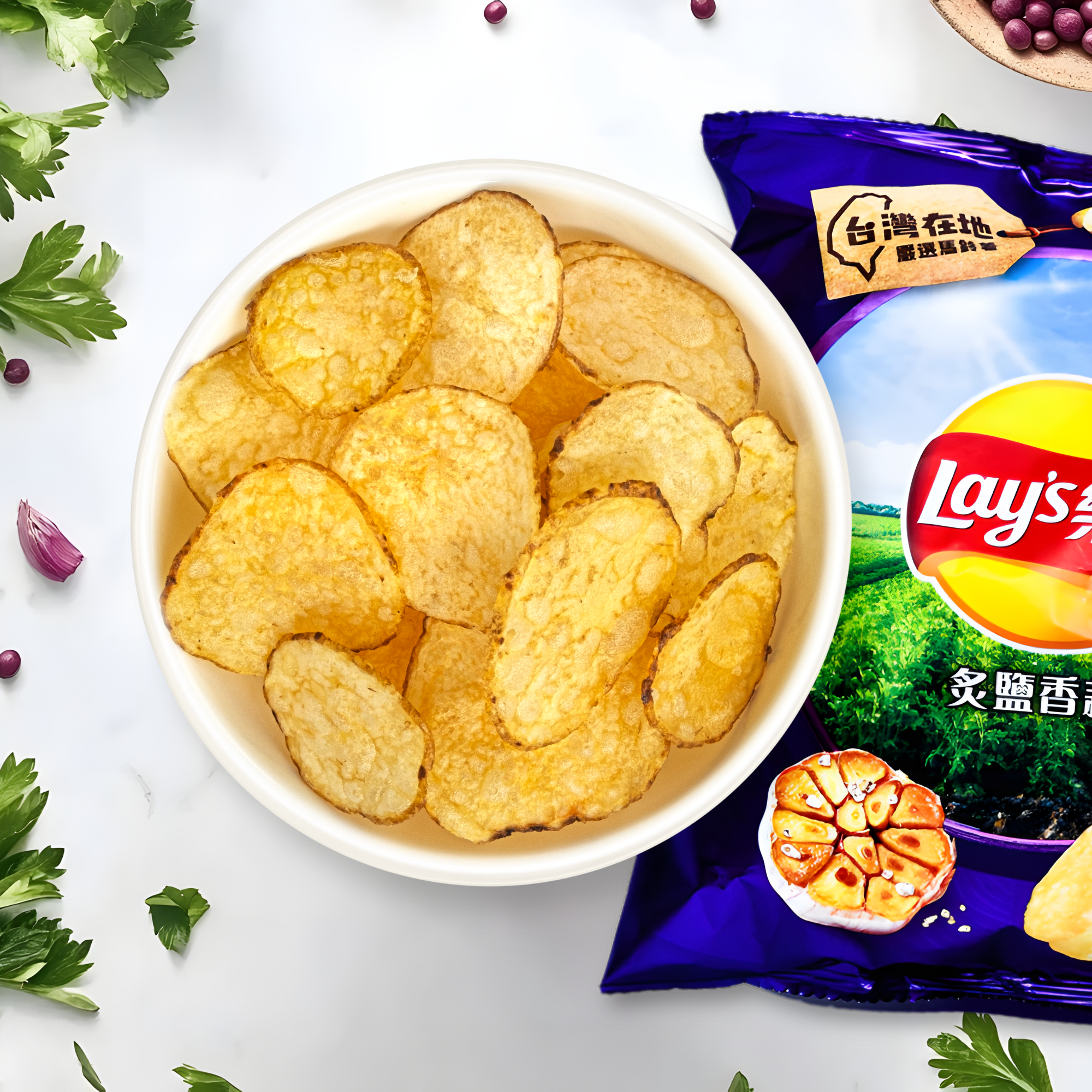 A bowl of crispy potato chips next to a bag of Lay's Roasted Garlic and Salt from Taiwan on a white surface, garnished with parsley and garlic cloves, conveying a savory snack vibe.