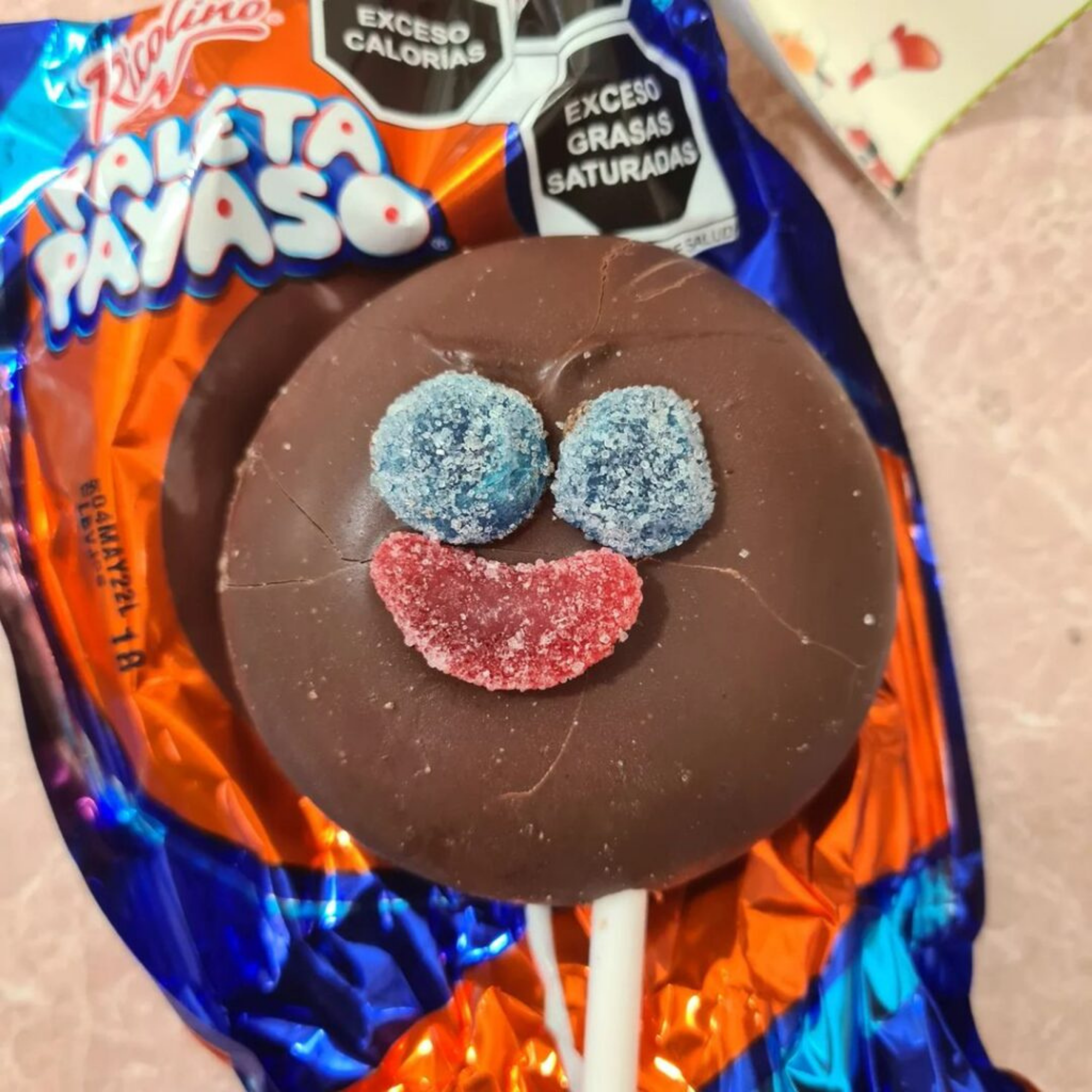 Paleta Payaso Chocolate lollipop with a clown face, featuring blue sugar eyes and a red sugar mouth. It's on colorful, vibrant candy packaging. Mexican candy available to buy online.