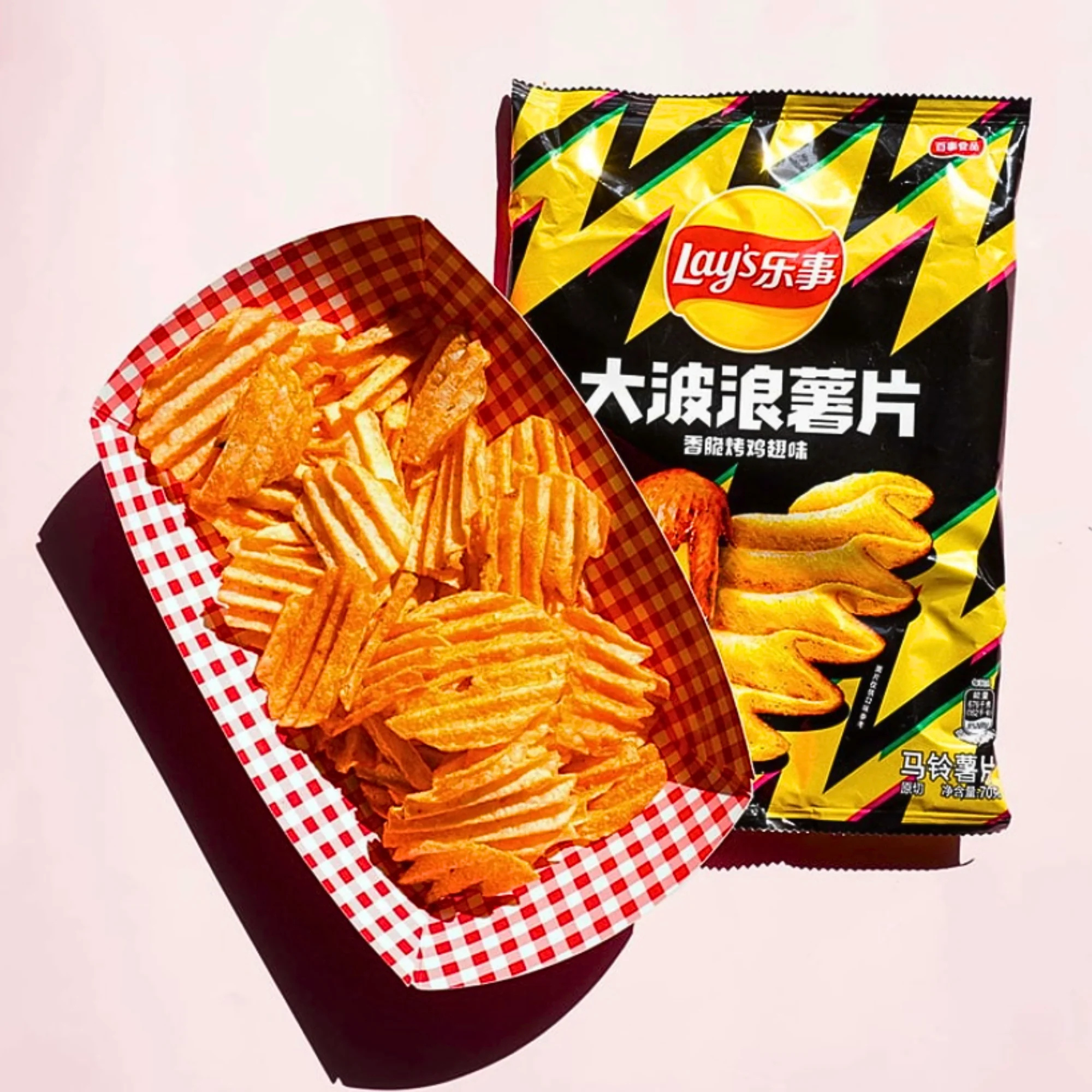 A vibrant red checkered basket filled with wavy, golden potato chips is placed beside a colorful Roasted Chicken Wing Asian Lay’s packaging. 