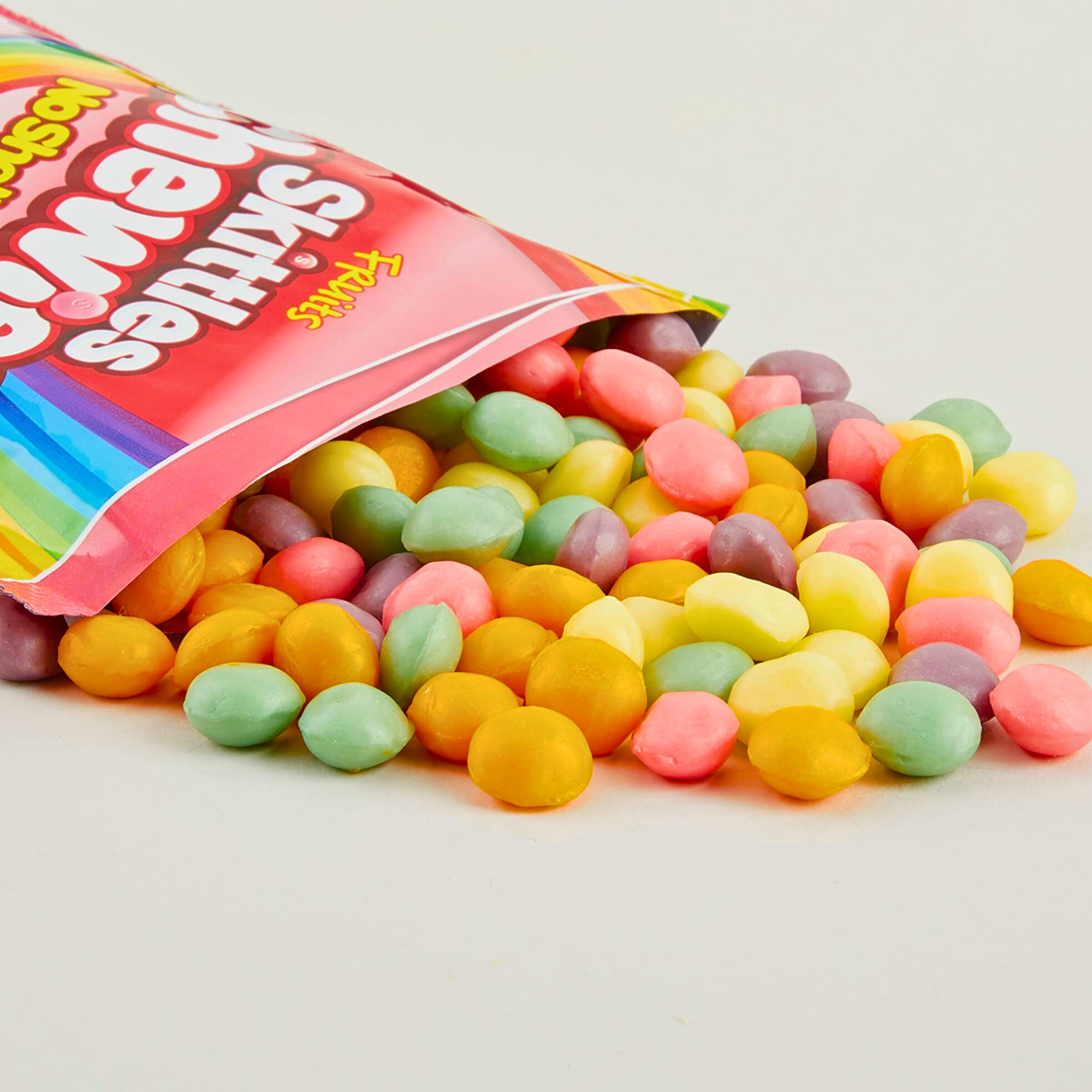 A colorful bag of Skittles Chewies 'No Shell' candy bag from the UK is spilling onto a white surface, showcasing vibrant candies in pink, yellow, green, orange and purple hues.
