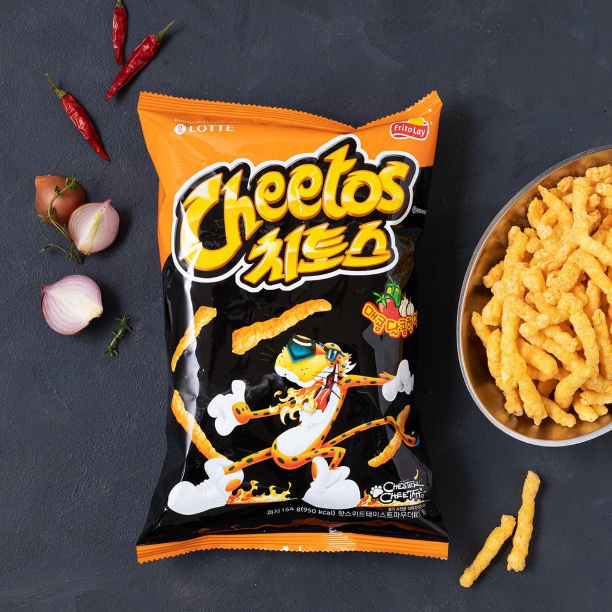 A black and orange bag of Korean Cheetos with a cartoon cheetah is next to a silver bowl filled with the crunchy cheetos snacks, surrounded by peppers and onions.