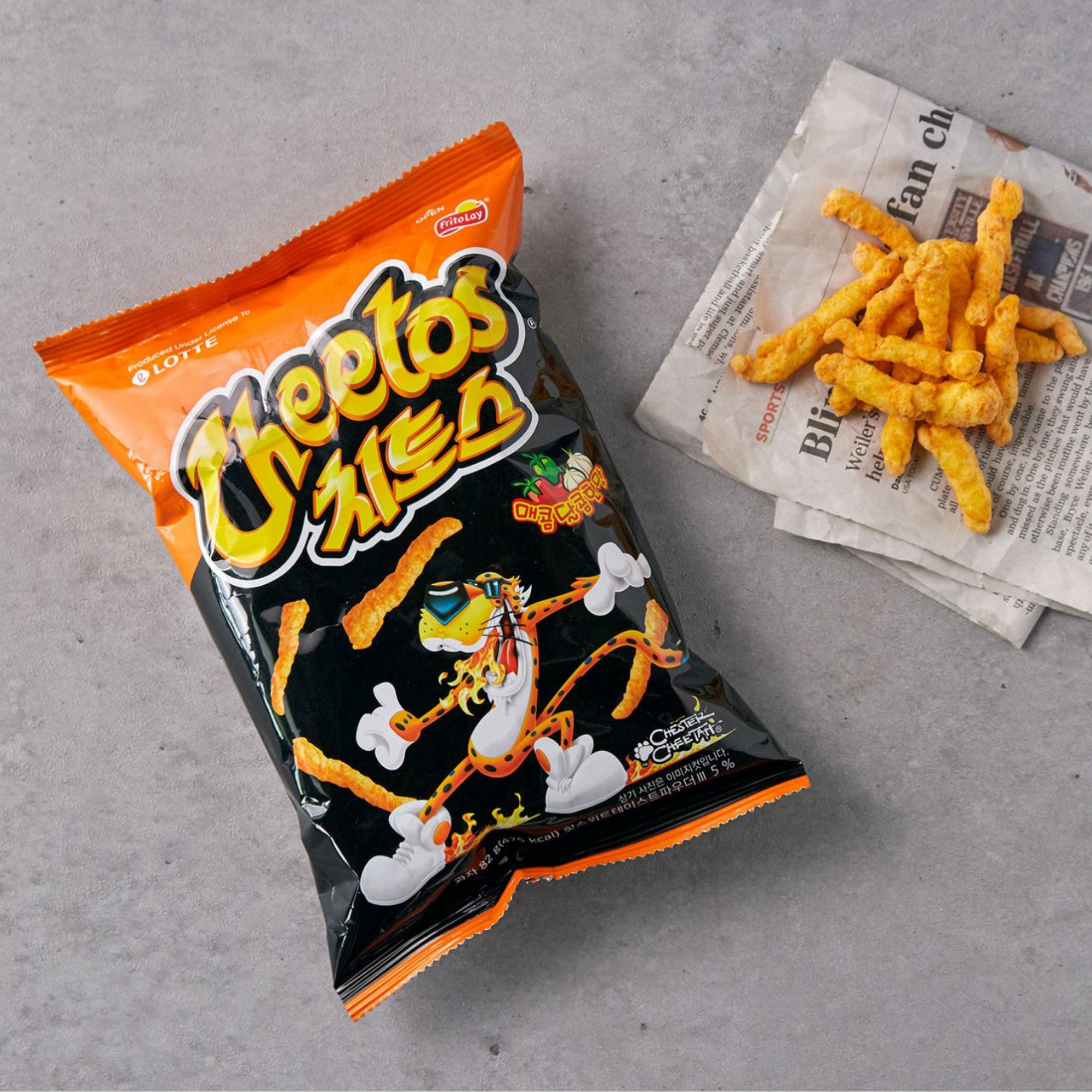 Bag of Korean Sweet and Spicy Cheetos with Korean text on a gray surface, next to crispy cheetos pieces on a newspaper.