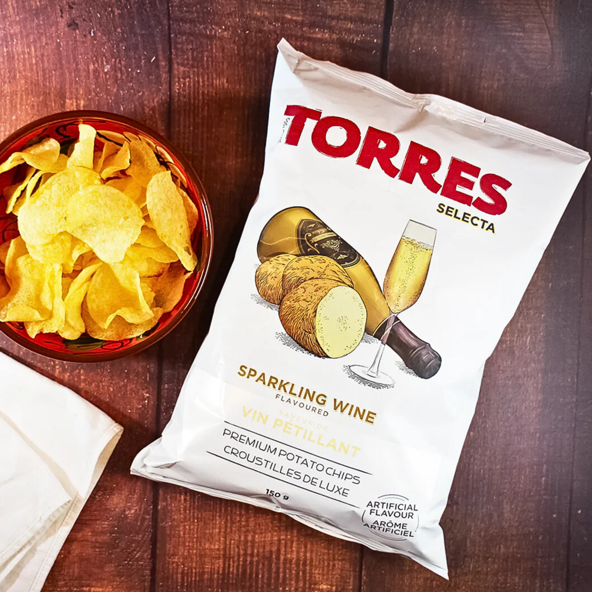 A white bag of Torres Selecta sparkling wine flavored potato chips from Spain lies on a wooden table. Next to it is a bowl filled with golden, crispy chips.