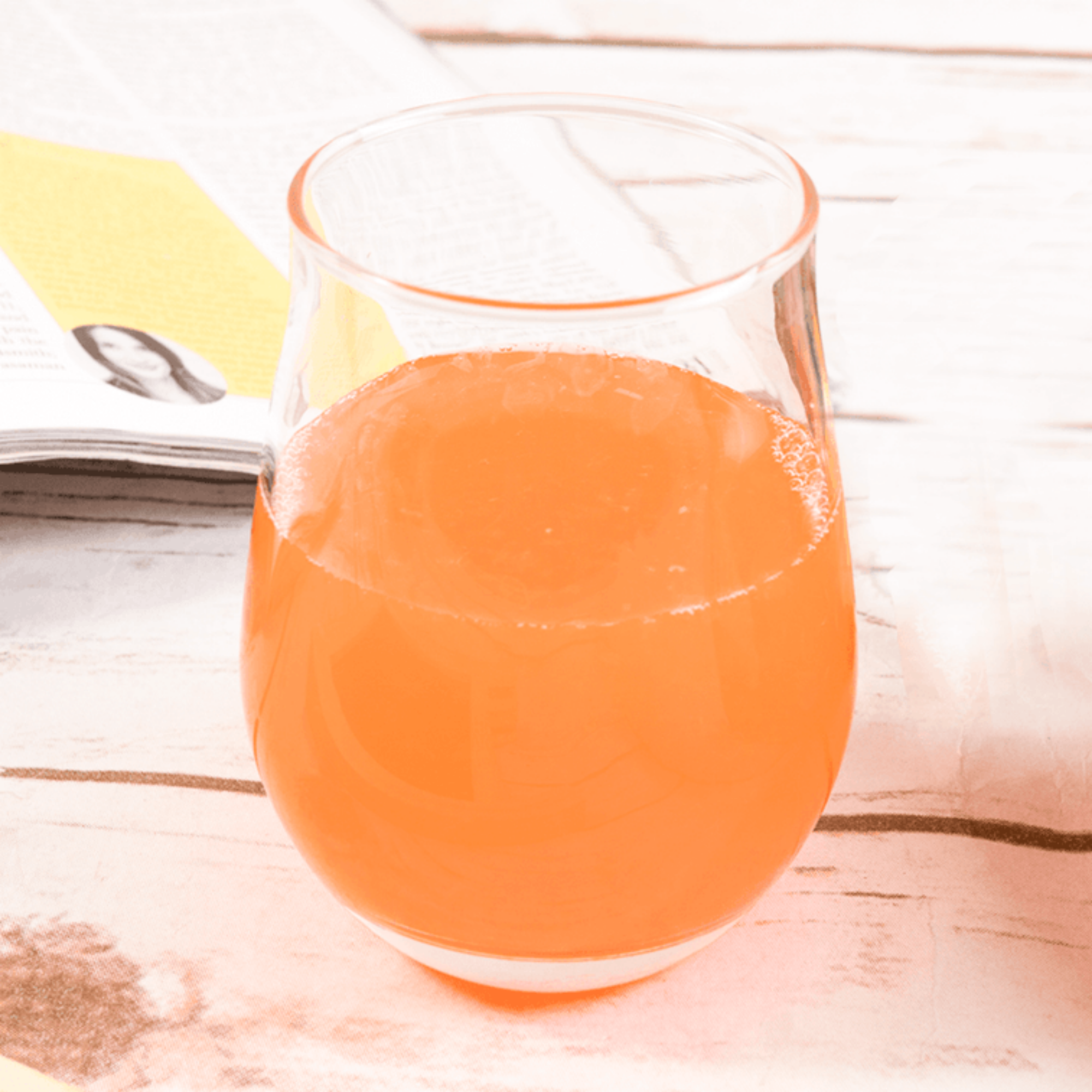 A clear glass filled with Tropical Punch Minute Maid juice from Taiwan sits on a wooden table beside an open magazine. 
